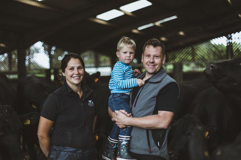 Man, woman and child on man's arms in cattle shed, looking at camera.